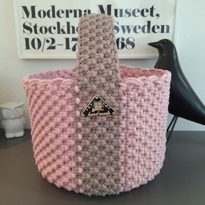Puede incluir: Una cesta redonda de ganchillo con un diseño de dos tonos en hilo rosa claro y color topo. La cesta tiene una sola asa gruesa y una etiqueta metálica triangular en la parte frontal con un gráfico de búho y el texto "hand made" grabado en oro.