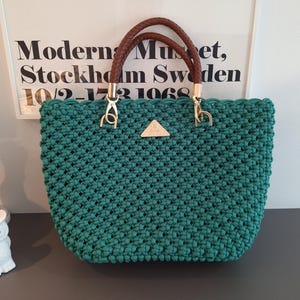 Könnte beinhalten: Eine dunkel-türkisfarbene Häkeltasche mit einem strukturierten Knotenmuster. Die Tasche hat zwei geflochtene braune Griffe, die mit goldfarbenen Metallbeschlägen befestigt sind. Eine kleine, dreieckige goldfarbene Metallplakette ist vorne mittig.