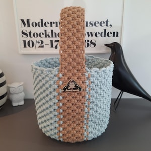 May include: A cylindrical macrame basket featuring a light blue body and a tan vertical centre stripe that extends into a single handle. A triangular metal tag with an owl graphic and the text "hand made" is attached to the centre stripe.