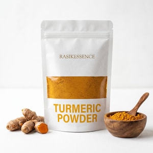 Peut inclure: Un sachet blanc debout étiqueté RASIKESSENCE TURMERIC POWDER avec une fenêtre transparente montrant de la poudre jaune-orange vif. À côté du sachet se trouvent plusieurs racines de curcuma et un petit bol en bois rempli de poudre.