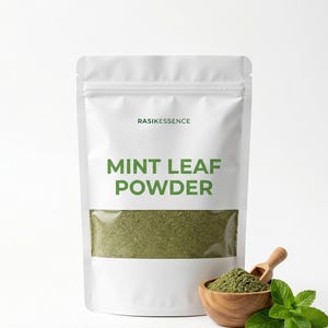 May include: A white stand-up pouch labeled RASIKESSENCE MINT LEAF POWDER with a clear window showing green powder. Next to the pouch is a small wooden bowl filled with the same green powder and a wooden scoop, accompanied by fresh green mint leaves.