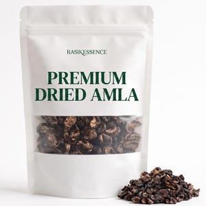 May include: A white stand-up pouch containing dark brown dried amla pieces. The label features the text RASIKESSENCE and PREMIUM DRIED AMLA in dark green lettering. A clear window reveals the contents, and a small pile of dried amla sits nearby.