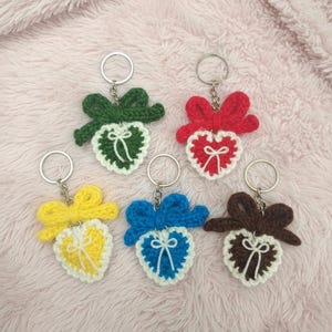 May include: A collection of six handmade crochet heart keychains in various colors: green, red, yellow, blue, and brown. Each keychain features a heart shape with a bow detail and a silver keyring.