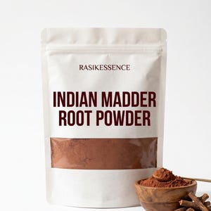 Könnte beinhalten: Ein weißer Standbeutel mit der Aufschrift RASIKESSENCE INDIAN MADDER ROOT POWDER und einem Sichtfenster, das braunes Pulver zeigt. Neben dem Beutel steht eine kleine Holzschale mit demselben braunen Pulver und einigen holzigen Wurzelstücken.