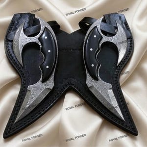 Op de afbeelding: Twee Damascus stalen messen met zwarte handvatten en scheden. De bladen hebben een uniek patroon en de handvatten zijn geaccentueerd met witte stippen. De messen worden gepresenteerd op een satijnen oppervlak, met de tekst "ROYAL FORGED" zichtbaar.
