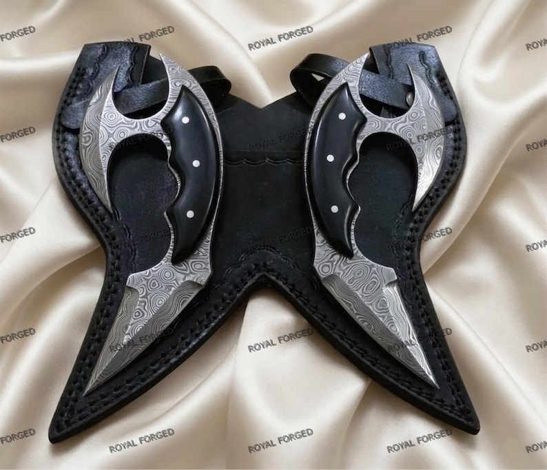 Op de afbeelding: Twee Damascus stalen messen met zwarte handvatten en scheden. De bladen hebben een uniek patroon en de handvatten zijn geaccentueerd met witte stippen. De messen worden gepresenteerd op een satijnen oppervlak, met de tekst "ROYAL FORGED" zichtbaar.