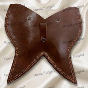 Op de afbeelding: Een bruine leren gorget in vlindervorm, met lichtbruine gestikte randen. Het heeft een kleine riem met een knoop. De achtergrond toont de tekst "Royal Forged".