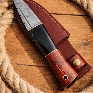 May include: A handcrafted knife with a patterned steel blade and a dark wooden handle. The knife is in a brown leather sheath with a brass button closure. A rope is coiled around the knife on a wooden surface.