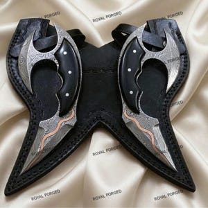 Op de afbeelding: Twee sierlijke, gebogen messen met patronen op de lemmeten en zwarte handvatten, elk in een zwarte leren schede. De lemmeten hebben een zilver- en koperkleurig ontwerp. De scheden zijn verbonden en hebben een riem. De woorden "ROYAL FORGED" staan op de scheden.