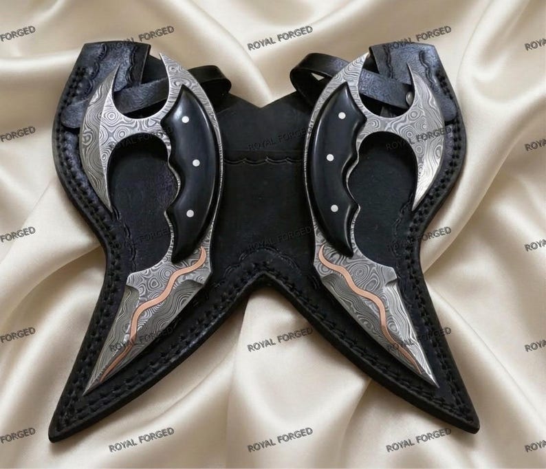 Op de afbeelding: Twee sierlijke, gebogen messen met patronen op de lemmeten en zwarte handvatten, elk in een zwarte leren schede. De lemmeten hebben een zilver- en koperkleurig ontwerp. De scheden zijn verbonden en hebben een riem. De woorden "ROYAL FORGED" staan op de scheden.