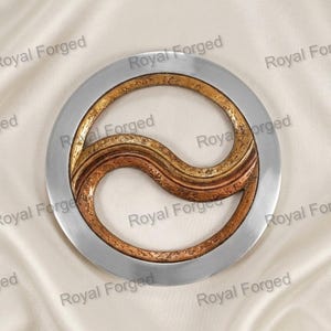 May include: A circular metal wall art piece featuring a smooth, polished silver outer ring surrounding an inner yin-yang shape. The inner design consists of two textured, curved sections in contrasting shades of bronze and copper metal.