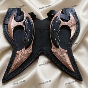 Op de afbeelding: Twee koperkleurige, gebogen messen met zwarte handvatten en een damaststalen patroon, rustend in een zwarte leren schede. De messen hebben een scherp, puntig lemmet. De schede heeft een riem om de messen vast te zetten. De woorden "ROYAL FORGED" staan op de afbeelding.