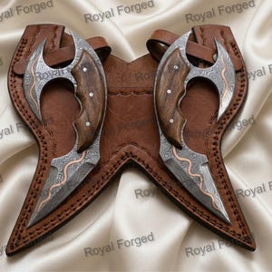 May include: Two curved knives with Damascus steel blades featuring wavy patterns and copper accents, paired with dark brown wooden handles. The knives are secured in a custom-fitted, stitched brown leather sheath with a symmetrical design.