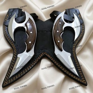 Op de afbeelding: Twee zilveren en houten messen met een zwarte leren schede. De messen hebben een gebogen lemmetontwerp en zitten in een zwarte leren schede met decoratieve stiksels. De afbeelding staat op een satijnen achtergrond.