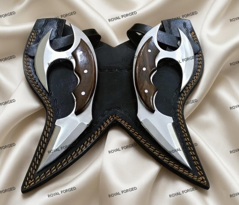 Op de afbeelding: Twee zilveren en houten messen met een zwarte leren schede. De messen hebben een gebogen lemmetontwerp en zitten in een zwarte leren schede met decoratieve stiksels. De afbeelding staat op een satijnen achtergrond.