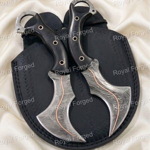 May include: Two curved, Damascus steel knives with black handles and copper accents, resting on a black leather sheath. The blades have a unique pattern, and the handles feature a finger ring. The text "Royal Forged" is visible.