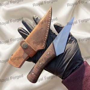 Op de afbeelding: Een handgemaakt jachtmes met een houten handvat en een roestvrijstalen lemmet, gepresenteerd met een bruine leren schede. Het mes heeft een full tang constructie en is ontworpen voor gebruik buitenshuis. De schede heeft een knoopsluiting.