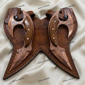 Op de afbeelding: Twee koperkleurige, gebogen messen met houten handvatten en decoratieve klinknagels, genesteld in een bruin lederen foedraal. De messen hebben een uniek patroon. De foedraal is gestikt en heeft een riem- en knoopsluiting. De woorden "ROYAL FORGED" staan op de foedraal.