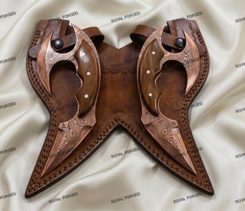 Op de afbeelding: Twee koperkleurige, gebogen messen met houten handvatten en decoratieve klinknagels, genesteld in een bruin lederen foedraal. De messen hebben een uniek patroon. De foedraal is gestikt en heeft een riem- en knoopsluiting. De woorden "ROYAL FORGED" staan op de foedraal.