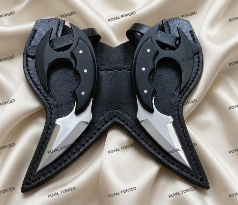Op de afbeelding: Twee zwarte en zilveren messen met gebogen lemmeten en zwarte handvatten, elk met drie witte stippen. De messen zitten in een zwart leren foedraal met de tekst "ROYAL FORGED".