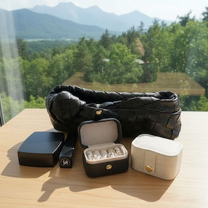 May include: A collection of black and white accessories, including a woven leather clutch, a small black jewelry box with rings, a white jewelry box, and a black compact. The items are arranged on a light-colored surface.