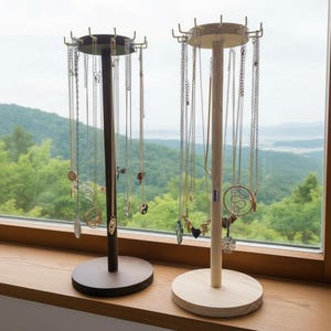 Puede incluir: Dos expositores de joyería de madera con múltiples collares colgando. Uno es de color marrón oscuro, el otro de madera clara. Los expositores tienen una base circular y una parte superior con ganchos para sujetar los collares. El fondo es una ventana con vistas a un paisaje verde.