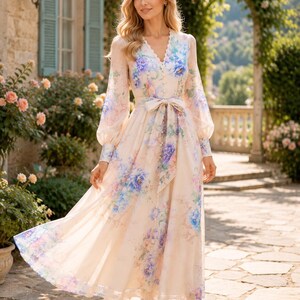 Puede incluir: Un vestido de manga larga hasta el suelo en color crema claro con un estampado floral vibrante en tonos azules, morados y verdes. El vestido presenta un escote en V, una parte delantera abotonada y un cintur&oacute;n de tela a juego en la cintura.