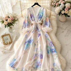 Puede incluir: Un vestido midi de manga larga en color crema claro con un estampado floral vibrante en tonos azul, morado y melocot&oacute;n. El vestido tiene un escote en V, cierre frontal de botones y un lazo de tela en la cintura. Las mangas son abullonadas y fruncidas en los pu&ntilde;os, y la falda tiene una silueta fluida en forma de A.