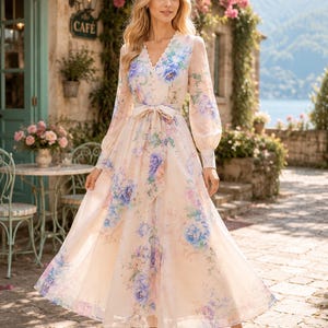 Puede incluir: Un vestido de manga larga y largo hasta el suelo en color crema claro con un vibrante estampado floral en tonos azules, morados y verdes. El vestido tiene un escote en V, cierre frontal de botones y un cintur&oacute;n de tela a juego.