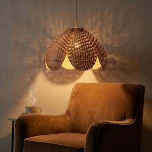 May include: A woven rattan pendant light fixture with a flower-like shape hangs above a brown velvet armchair. The light casts a warm glow, illuminating the interior. A small side table with a cup of tea completes the cozy scene.