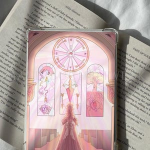 Könnte beinhalten: Eine transparente Tablet-Hülle mit einer rosa Illustration einer Person in einem langen Kleid, die in einem kirchenartigen Innenraum vor Buntglasfenstern, einer großen runden Uhr, Pfeilen, Spielkarten und einem Baum mit Wurzeln steht.