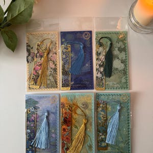 Könnte beinhalten: Sechs dekorative Lesezeichen in transparenten Kunststoffhüllen. Jedes Lesezeichen hat ein einzigartiges Design mit floralen oder künstlerischen Themen, eine Quaste und einen goldfarbenen Metallakzent. Die Farbpalette umfasst Blau-, Grün- und Gelbtöne.