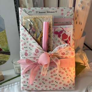 Peut inclure: Un coffret cadeau enveloppé dans du papier imprimé de cerises, noué avec un ruban et un nœud roses. La boîte contient un marque-page, des autocollants et un stylo rose. Le texte "Summer Romance" est imprimé sur le dessus.