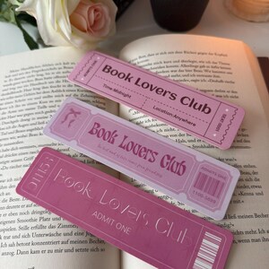 以下が含まれることがあります： 「Book Lovers Club」の文字が入った、チケット型のピンク色のブックマークが3つ。ブックマークには「Admit One」、「Time: Midnight」、バーコードなどの詳細が記載されています。開いた本の上に配置されています。