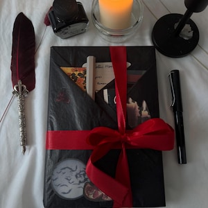 Könnte beinhalten: Ein Geschenkset zum Thema Gothic mit einer schwarzen Verpackung, die mit einem roten Band gebunden ist. Das Set enthält eine Federkiel, eine Tintenflasche, eine Kerze und einen Stift. Das Paket hat einen Aufkleber mit Mond und Fledermäusen.