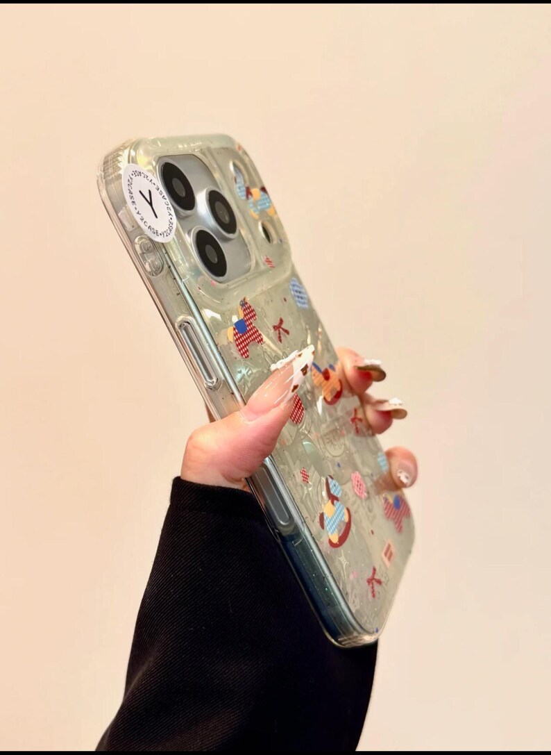 Puede incluir: Funda transparente para tel&eacute;fono con dise&ntilde;os coloridos y caprichosos. La funda presenta varias formas y patrones, incluyendo un instrumento musical y un dise&ntilde;o de punto de cruz. Una pegatina blanca con una "Y" negra es visible.