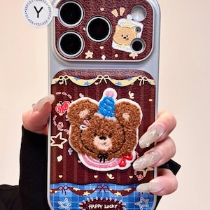 Puede incluir: Funda de teléfono decorativa con un oso de peluche marrón con un gorro de fiesta azul y un lazo rojo. La funda tiene un fondo burdeos con estrellas y el texto "HAPPY LUCKY". La funda tiene un marco plateado y un diseño de oso.