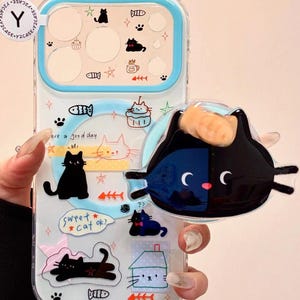 Puede incluir: Funda transparente para teléfono con borde azul claro, con ilustraciones y texto con temática de gatos. Un charm 3D de gato negro y naranja está adjunto. La funda tiene varios gráficos de gatos y espinas de pescado.