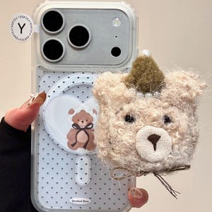 May include: A clear phone case with a teddy bear design. The case features a fluffy, light brown teddy bear head with a small crown and pearl accents. The case also has a heart-shaped design with a teddy bear illustration and small black hearts.