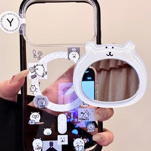 May include: Clear phone case with a black border, featuring cartoon bear stickers and a white bear-shaped mirror. The case has a transparent back, showcasing the phone's design. The mirror is attached to the case, adding a functional and cute element.