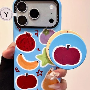 May include: A light blue phone case with a fruit design, including a red apple, banana, and pomegranate. A matching blue pop socket with a red apple design is also visible. The case has a protective bumper around the camera.