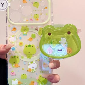 Puede incluir: Una funda transparente para teléfono con borde verde claro, con un patrón repetido de ranas, peces y estrellas de dibujos animados. Un dije en forma de rana está adjunto. La funda tiene texto japonés.