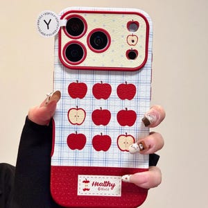 Könnte beinhalten: Eine Handyhülle mit einem rot-weißen Apfelmuster. Die Hülle hat einen roten Rand und ein blau-weißes Karomuster. Das Design umfasst rote Apfelillustrationen und das Wort "Healthy".