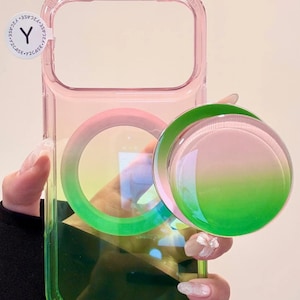 May include: A clear phone case with a pink and green gradient design. The case has a circular pop-up grip with a matching gradient. The case has a clear window for the phone's camera and a sticker with the letter "Y".