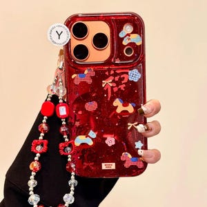 Puede incluir: Funda de tel&eacute;fono roja con un dise&ntilde;o decorativo con caballos, nubes y estrellas. La funda tiene una correa de mu&ntilde;eca a juego con cuentas rojas y transparentes. La funda tiene un acabado transparente y brillante.