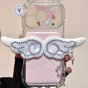 May include: Clear phone case with a pink interior and white fluffy angel wings embellished with rhinestones. The case has a clear back with decorative stickers and charms, including a star and a bell.