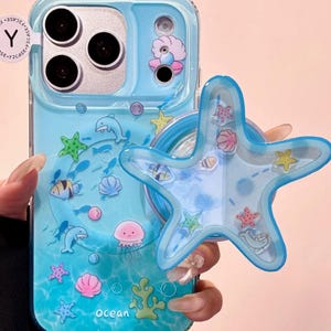 Puede incluir: Funda de teléfono azul claro con temática oceánica, con delfines, estrellas de mar y otras criaturas marinas. Incluye un pop-socket a juego con forma de estrella de mar. La palabra "ocean" está impresa en la funda.