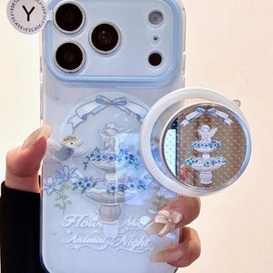 May include: A light blue phone case with a decorative design featuring a fountain, angel, and flowers. The case has a matching pop-socket with a similar design. The text "Flower, Animal, Night" is visible.
