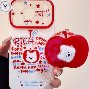 Puede incluir: Funda de teléfono blanca con borde rojo y agarre en forma de manzana roja. La funda presenta una ilustración de oso y el texto "RICH", "Money", "Luck" y "Have a nice day!"