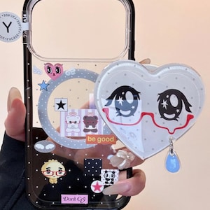 May include: Clear phone case with a black border, featuring a heart-shaped charm with a cartoon face and a blue teardrop. The case has various cute graphics, including stars, bears, and the text "be good" and "Don't Cry".